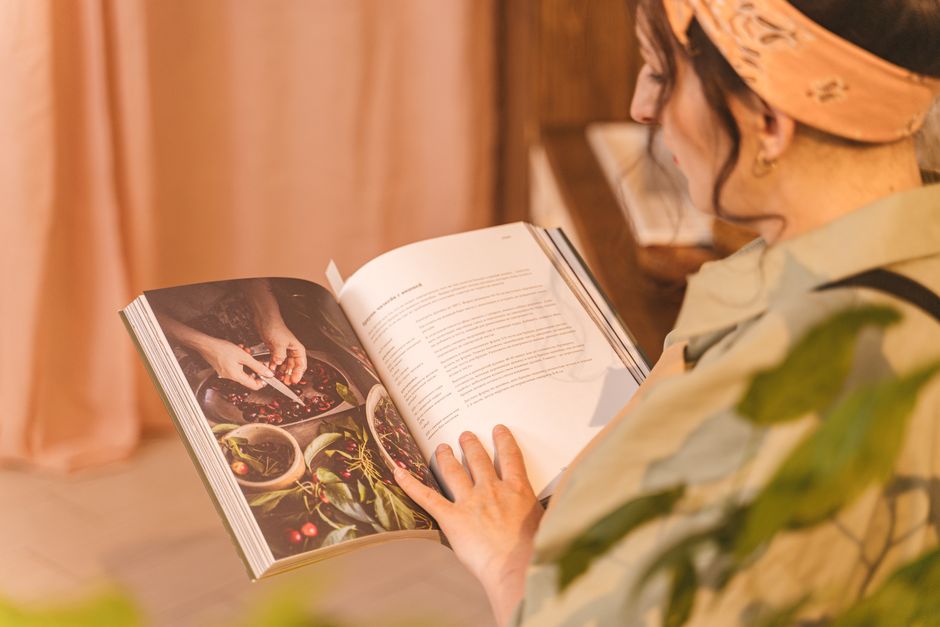 A woman reading a cookbook with colorful images and recipes in a warm indoor setting.